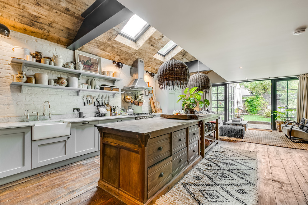 Photo of a mid-sized tropical galley open plan kitchen in London with a farmhouse sink, shaker cabinets, grey cabinets, wood benchtops, white splashback, brick splashback, medium hardwood floors, with island, brown floor and brown benchtop.