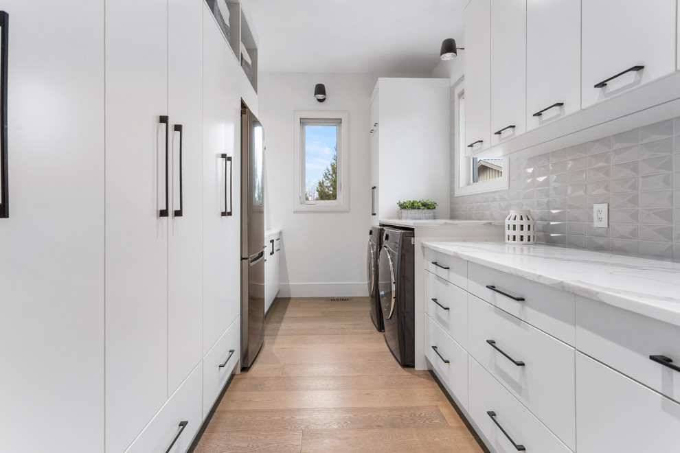 This is an example of a mid-sized modern galley utility room in Vancouver with an undermount sink, flat-panel cabinets, white cabinets, quartzite benchtops, grey splashback, porcelain splashback, white walls, medium hardwood floors, a side-by-side washer and dryer, brown floor and multi-coloured benchtop.