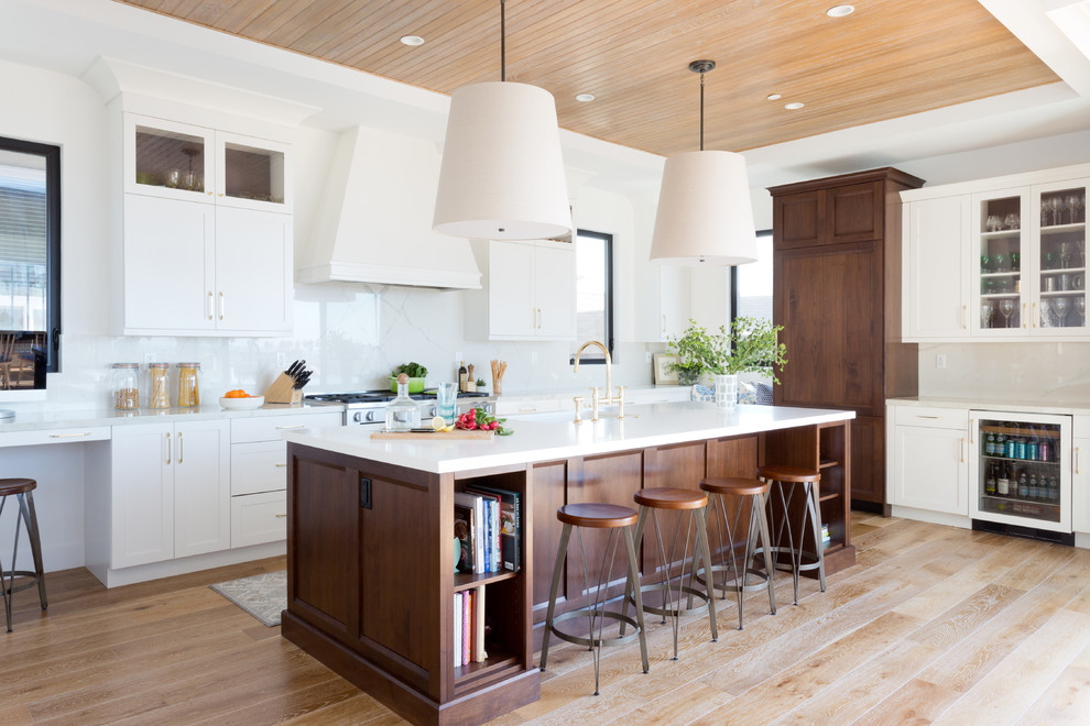 This is an example of a transitional l-shaped open plan kitchen in Los Angeles with recessed-panel cabinets, white cabinets, white splashback, marble splashback, panelled appliances, light hardwood floors, with island and beige floor.
