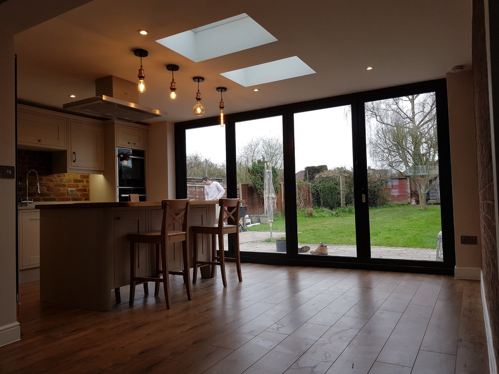 Mid-sized modern single-wall eat-in kitchen in Surrey with an integrated sink, louvered cabinets, beige cabinets, wood benchtops, red splashback, stone tile splashback, panelled appliances, laminate floors, with island, brown floor and brown benchtop.