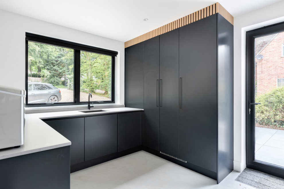 Photo of an expansive modern l-shaped utility room in Hertfordshire with an integrated sink, flat-panel cabinets, black cabinets, quartzite benchtops, white walls, porcelain floors, a concealed washer and dryer, grey floor and grey benchtop.
