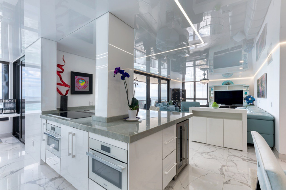 Mid-sized beach style galley open plan kitchen in Miami with flat-panel cabinets, white cabinets, marble benchtops, white splashback, white appliances, marble floors, with island, white floor, grey benchtop and wallpaper.