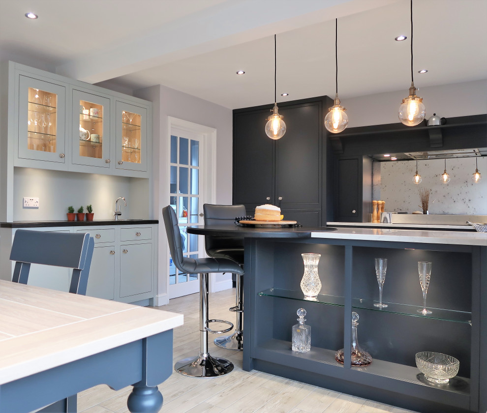 Modern inredning av ett mellanstort vit vitt kök, med en dubbel diskho, släta luckor, blå skåp, bänkskiva i kvartsit, spegel som stänkskydd, svarta vitvaror, klinkergolv i keramik, en halv köksö och brunt golv