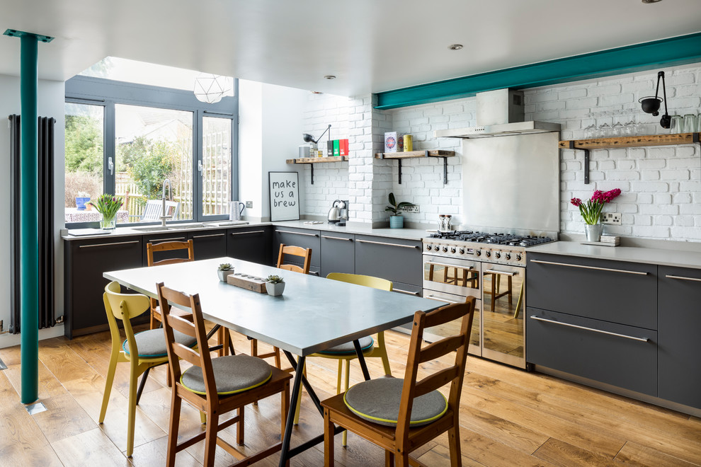 This is an example of an industrial l-shaped eat-in kitchen in London with a double-bowl sink, flat-panel cabinets, grey cabinets, concrete benchtops, white splashback, brick splashback, stainless steel appliances, medium hardwood floors and no island.
