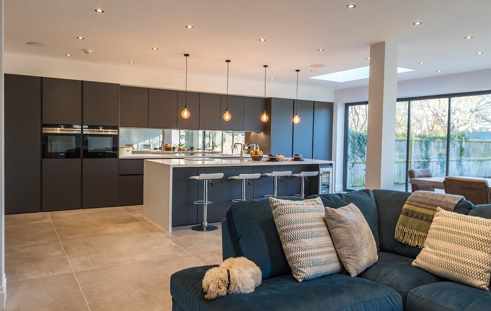 Photo of a large modern single-wall open plan kitchen in Surrey with a drop-in sink, flat-panel cabinets, grey cabinets, solid surface benchtops, metallic splashback, glass sheet splashback, black appliances, ceramic floors, with island and grey floor.