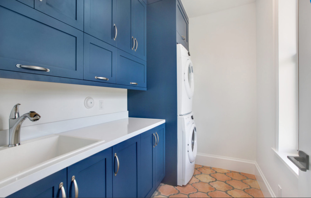 This is an example of a mid-sized transitional galley dedicated laundry room in Miami with a drop-in sink, shaker cabinets, blue cabinets, solid surface benchtops, white walls, terra-cotta floors and a stacked washer and dryer.
