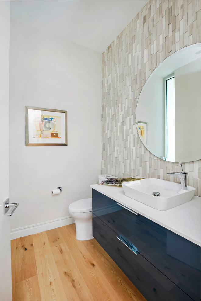 This is an example of a mid-sized contemporary powder room in Tampa with flat-panel cabinets, blue cabinets, a two-piece toilet, gray tile, stone tile, white walls, light hardwood floors, a vessel sink, engineered quartz benchtops, brown floor and white benchtops.