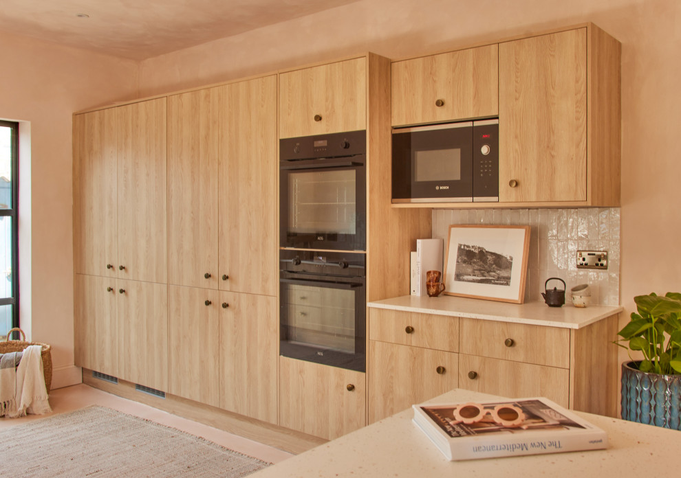 Inspiration for a large mediterranean l-shaped eat-in kitchen in Cornwall with an undermount sink, flat-panel cabinets, light wood cabinets, solid surface benchtops, beige splashback, ceramic splashback, black appliances, concrete floors, a peninsula, pink floor and beige benchtop.