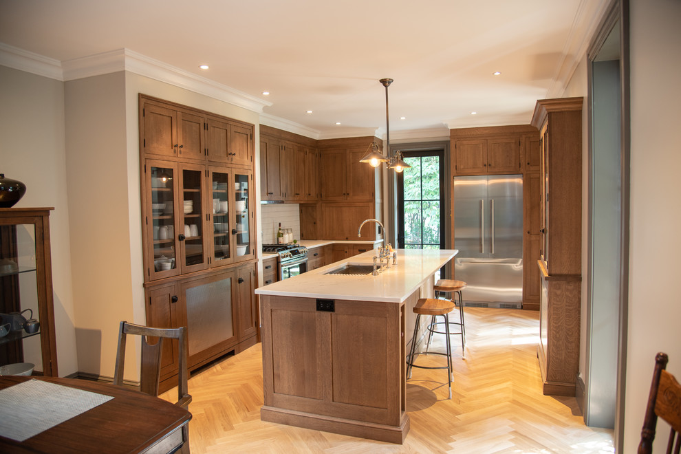 This is an example of a mid-sized arts and crafts l-shaped eat-in kitchen in Philadelphia with an undermount sink, shaker cabinets, medium wood cabinets, quartz benchtops, white splashback, ceramic splashback, stainless steel appliances, light hardwood floors, with island and white benchtop.