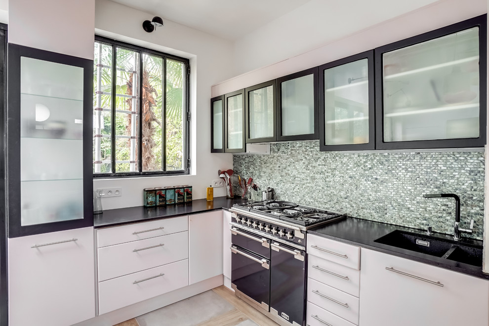 Photo of a large industrial l-shaped kitchen in Paris with flat-panel cabinets, mosaic tile splashback, black appliances, ceramic floors, black benchtop, a double-bowl sink, white cabinets and metallic splashback.