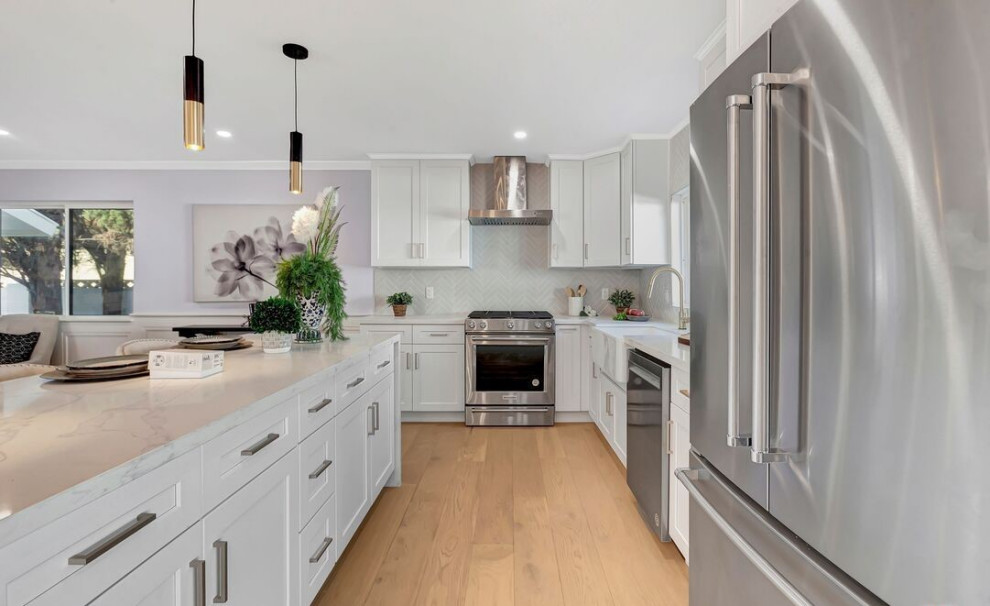 Photo of a contemporary l-shaped kitchen in Orange County with a farmhouse sink, shaker cabinets, white cabinets, marble benchtops, white splashback, ceramic splashback, stainless steel appliances, light hardwood floors, with island, brown floor and white benchtop.