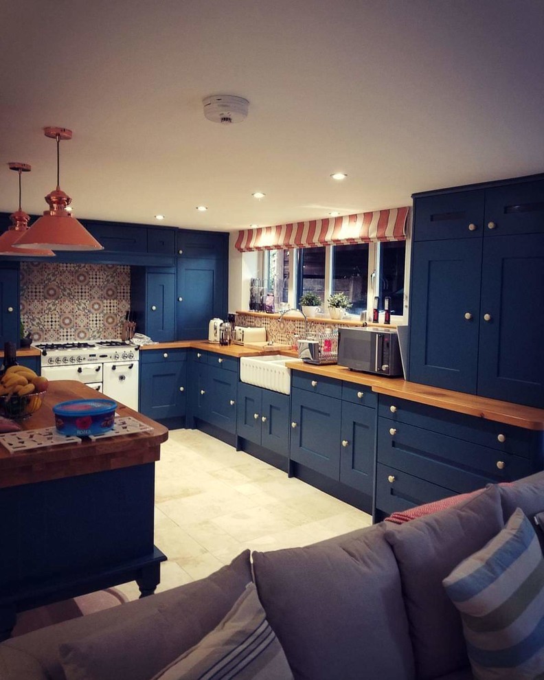 This is an example of a large eclectic l-shaped open plan kitchen in Baltimore with a farmhouse sink, blue cabinets, wood benchtops, multi-coloured splashback, mosaic tile splashback, white appliances, limestone floors, with island, beige floor, brown benchtop and shaker cabinets.