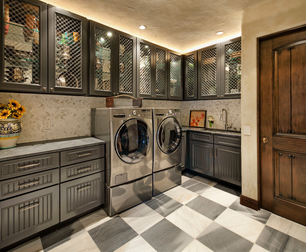 Design ideas for a large mediterranean l-shaped dedicated laundry room in Santa Barbara with an undermount sink, glass-front cabinets, grey cabinets, marble benchtops, beige walls, porcelain floors and a side-by-side washer and dryer.