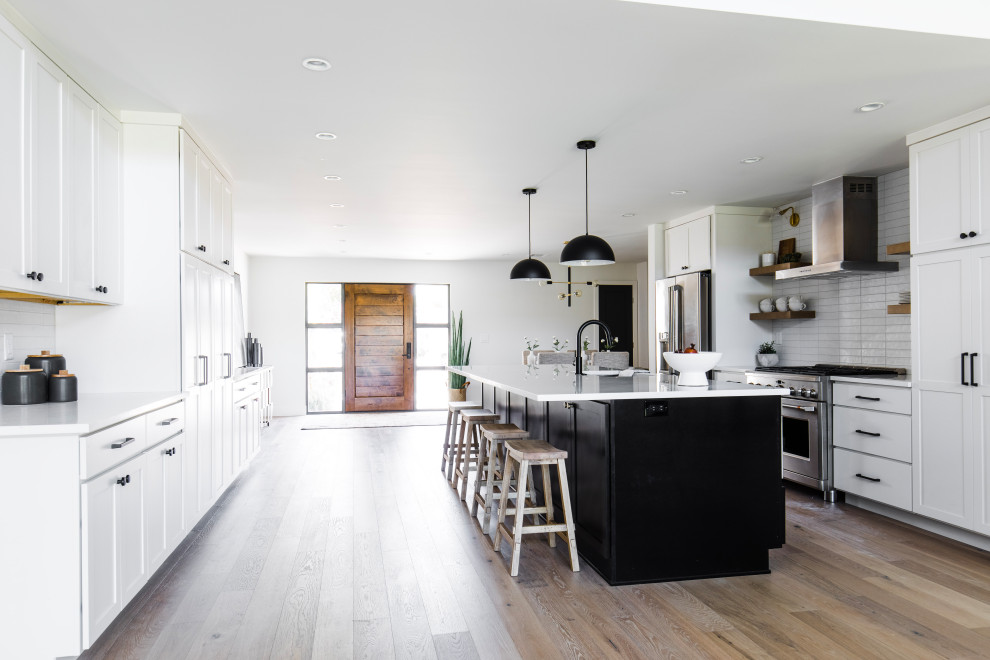 Open concept kitchen - large transitional galley medium tone wood floor open concept kitchen idea in Phoenix with a single-bowl sink, shaker cabinets, white cabinets, quartz countertops, white backsplash, ceramic backsplash, stainless steel appliances, an island and white countertops