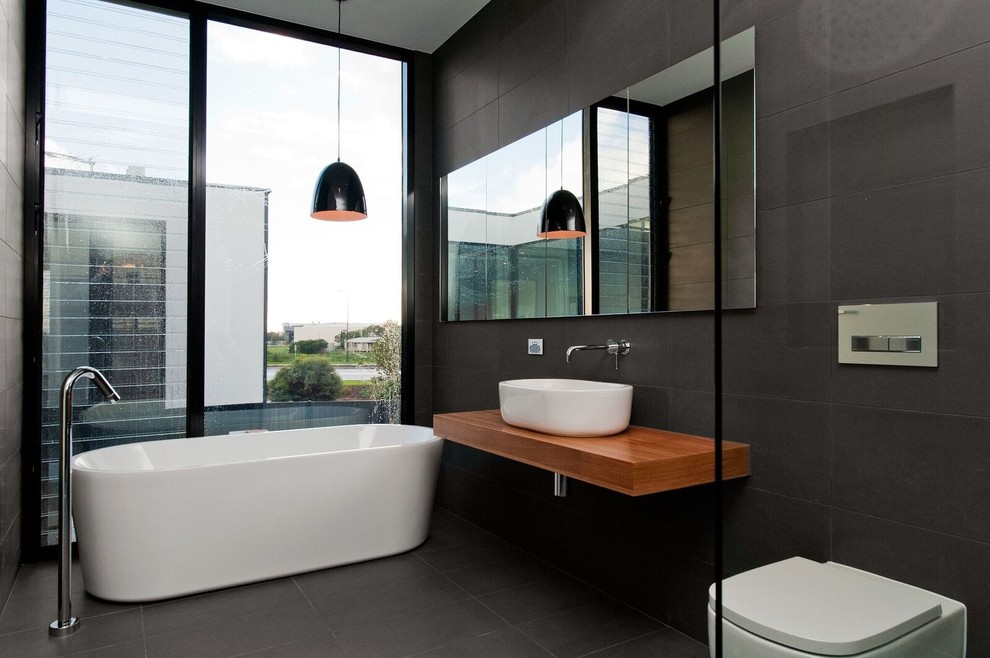 Inspiration for a mid-sized contemporary master bathroom in Adelaide with a freestanding tub, black tile, black walls, a vessel sink, medium wood cabinets, a one-piece toilet, cement tile, wood benchtops, black floor, an open shower and brown benchtops.