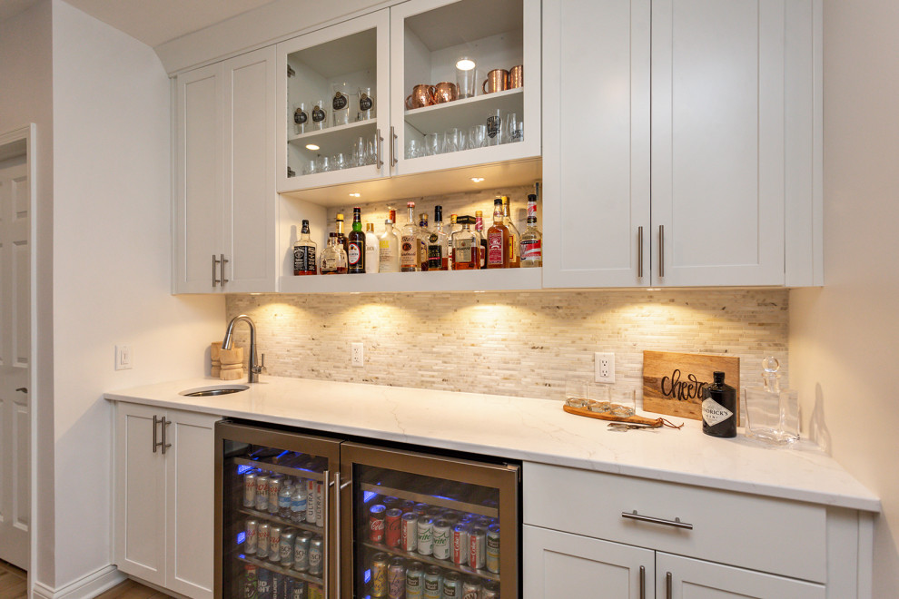 Photo of a large transitional single-wall wet bar in Other with an undermount sink, recessed-panel cabinets, white cabinets, quartz benchtops, white splashback, mosaic tile splashback, light hardwood floors, brown floor and white benchtop.