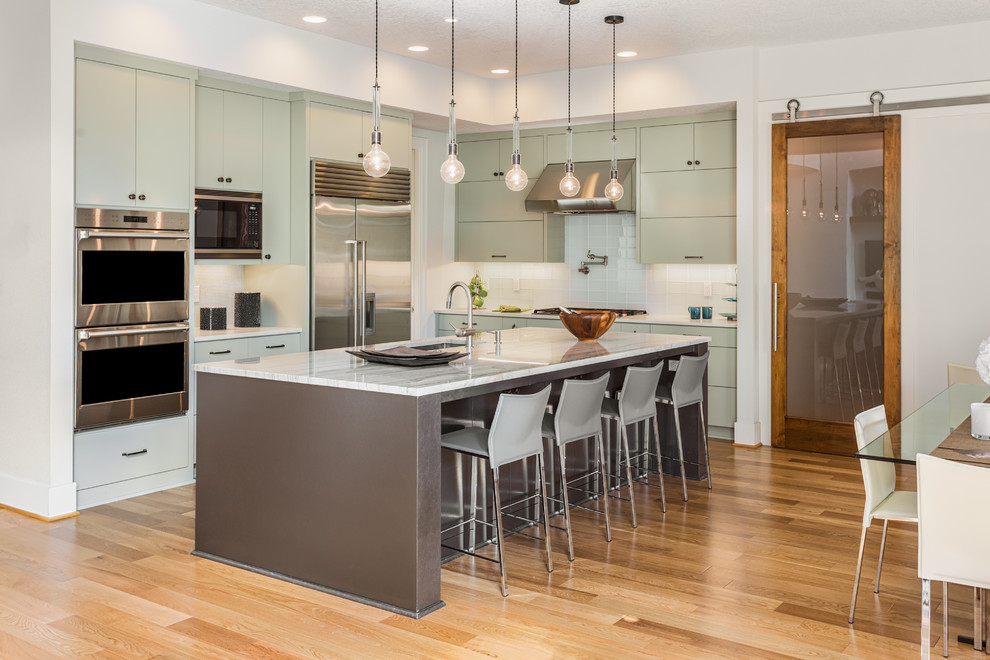 This is an example of a contemporary eat-in kitchen in DC Metro with an undermount sink, flat-panel cabinets, green cabinets, white splashback, stainless steel appliances, light hardwood floors, with island, beige floor and white benchtop.