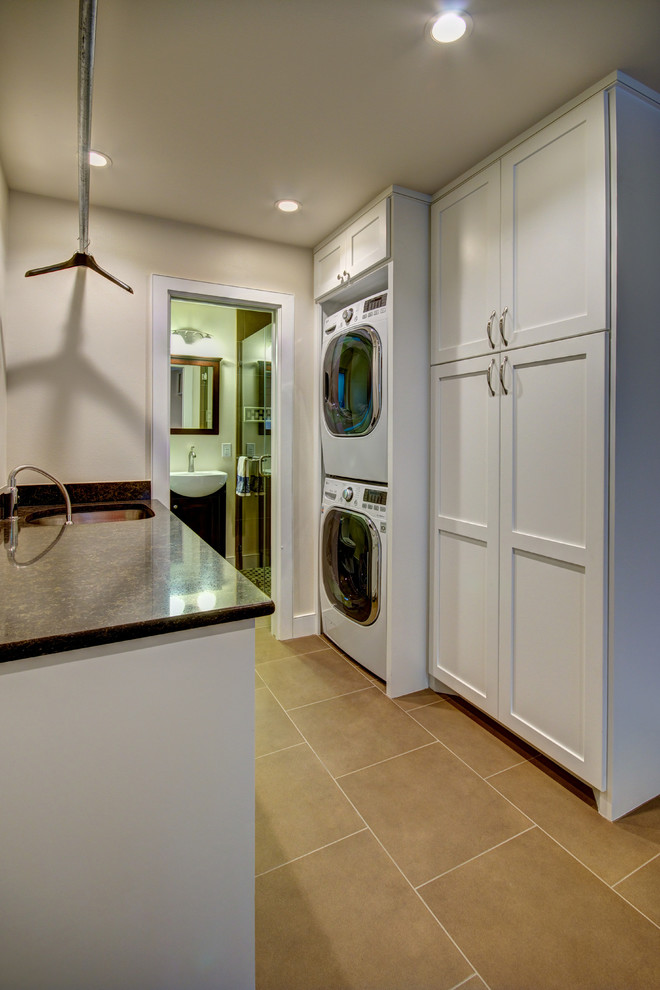 This is an example of a mid-sized transitional galley utility room in Austin with an undermount sink, shaker cabinets, white cabinets, granite benchtops, beige walls, limestone floors, a stacked washer and dryer and beige floor.
