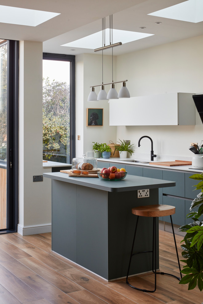 This is an example of a contemporary galley open plan kitchen in London with a drop-in sink, flat-panel cabinets, grey cabinets, white splashback, medium hardwood floors, with island and brown floor.