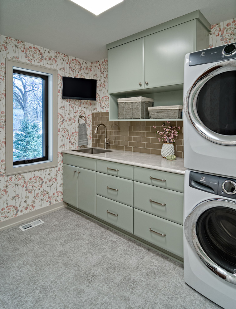 This is an example of a mid-sized transitional galley dedicated laundry room in Minneapolis with an undermount sink, flat-panel cabinets, green cabinets, laminate benchtops, multi-coloured walls, vinyl floors, a stacked washer and dryer, grey floor and grey benchtop.