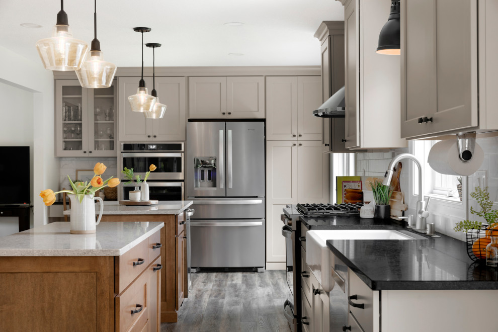 This is an example of a large transitional single-wall eat-in kitchen in Minneapolis with a farmhouse sink, shaker cabinets, grey cabinets, quartz benchtops, white splashback, ceramic splashback, stainless steel appliances, vinyl floors, multiple islands, brown floor and black benchtop.