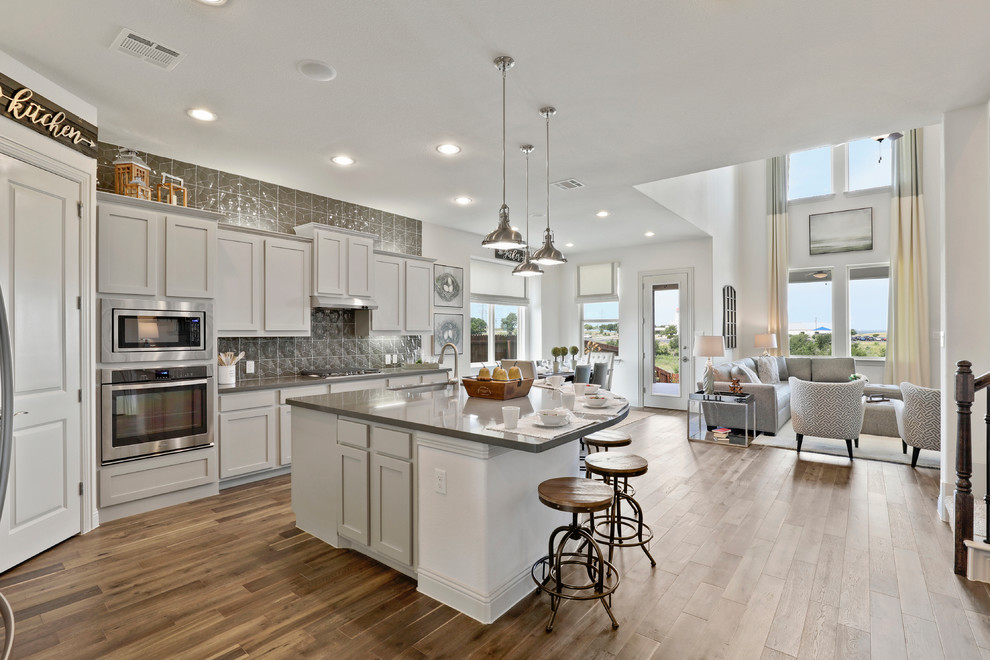 Photo of a large contemporary l-shaped eat-in kitchen in Dallas with a drop-in sink, shaker cabinets, grey cabinets, solid surface benchtops, grey splashback, glass sheet splashback, stainless steel appliances, dark hardwood floors, with island, brown floor and grey benchtop.