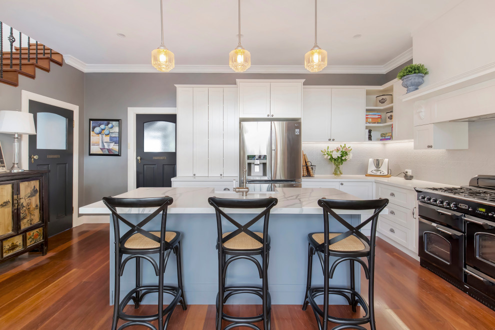 Mid-sized traditional l-shaped eat-in kitchen in Sydney with a farmhouse sink, shaker cabinets, white cabinets, quartz benchtops, grey splashback, metal splashback, black appliances, medium hardwood floors, with island, brown floor and multi-coloured benchtop.