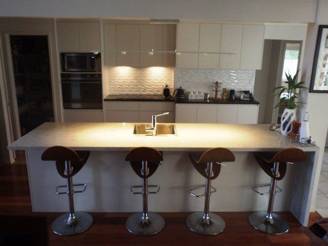 Mid-sized modern galley eat-in kitchen in Sunshine Coast with a drop-in sink, flat-panel cabinets, white cabinets, quartz benchtops, white splashback, ceramic splashback, black appliances and with island.