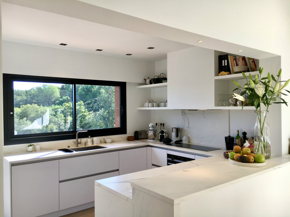 Mid-sized contemporary u-shaped eat-in kitchen in Other with a single-bowl sink, beaded inset cabinets, white cabinets, solid surface benchtops, white splashback, panelled appliances, limestone floors, beige floor, white benchtop and a peninsula.