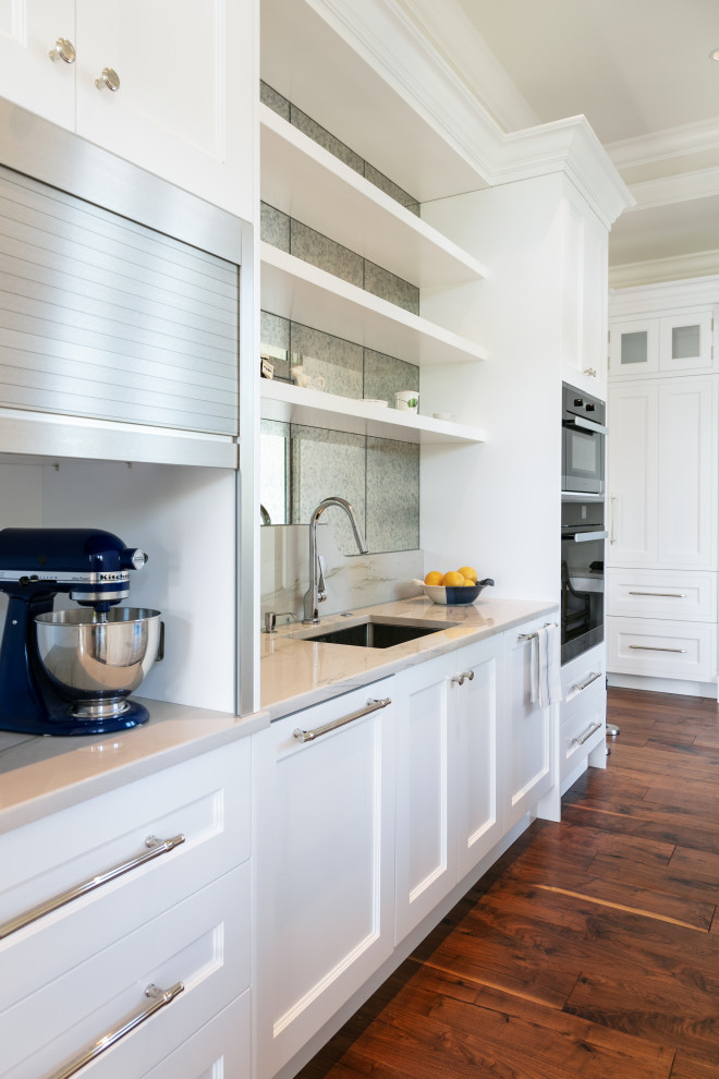 Photo of a small beach style galley kitchen pantry in Tampa with an integrated sink, raised-panel cabinets, white cabinets, granite benchtops, metallic splashback, mirror splashback, black appliances, dark hardwood floors, no island, brown floor and white benchtop.