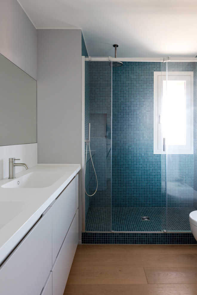 This is an example of a large contemporary 3/4 bathroom in Milan with beaded inset cabinets, light wood cabinets, blue tile, mosaic tile, grey walls, light hardwood floors, an integrated sink, a sliding shower screen and white benchtops.