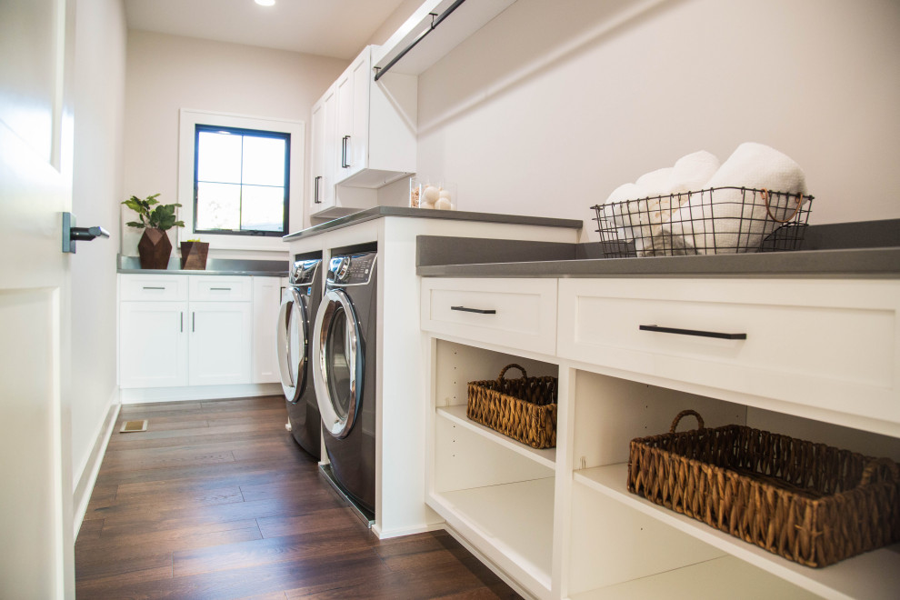 Exempel på en stor modern grå l-formad grått tvättstuga enbart för tvätt, med en undermonterad diskho, luckor med infälld panel, vita skåp, granitbänkskiva, vita väggar, mellanmörkt trägolv, en tvättmaskin och torktumlare bredvid varandra och brunt golv
