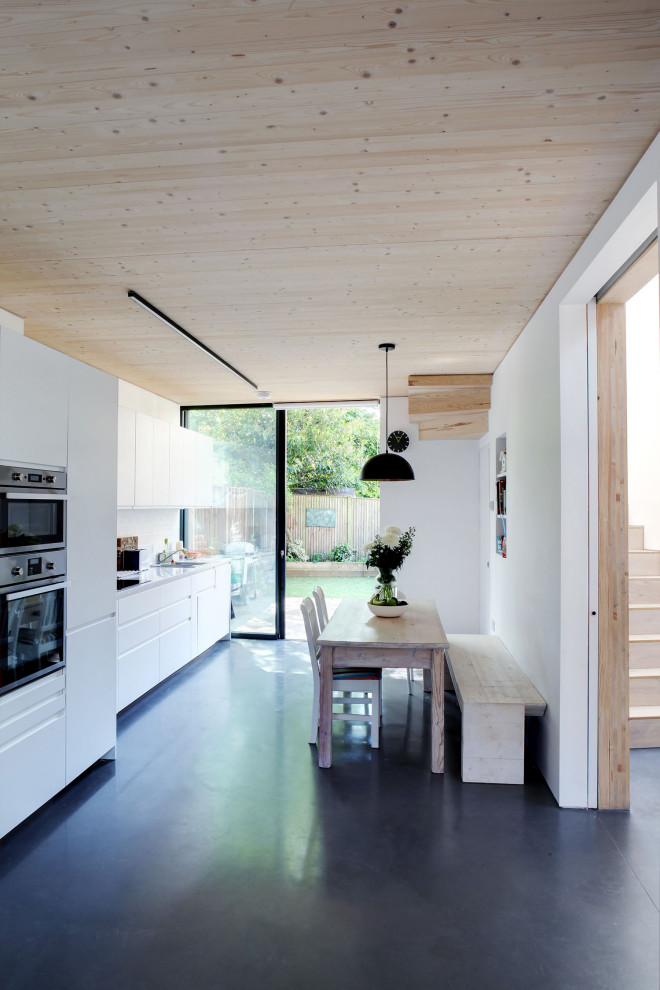 Photo of a mid-sized contemporary galley eat-in kitchen in London with an undermount sink, flat-panel cabinets, white cabinets, white splashback, panelled appliances, no island, black floor, white benchtop and wood.