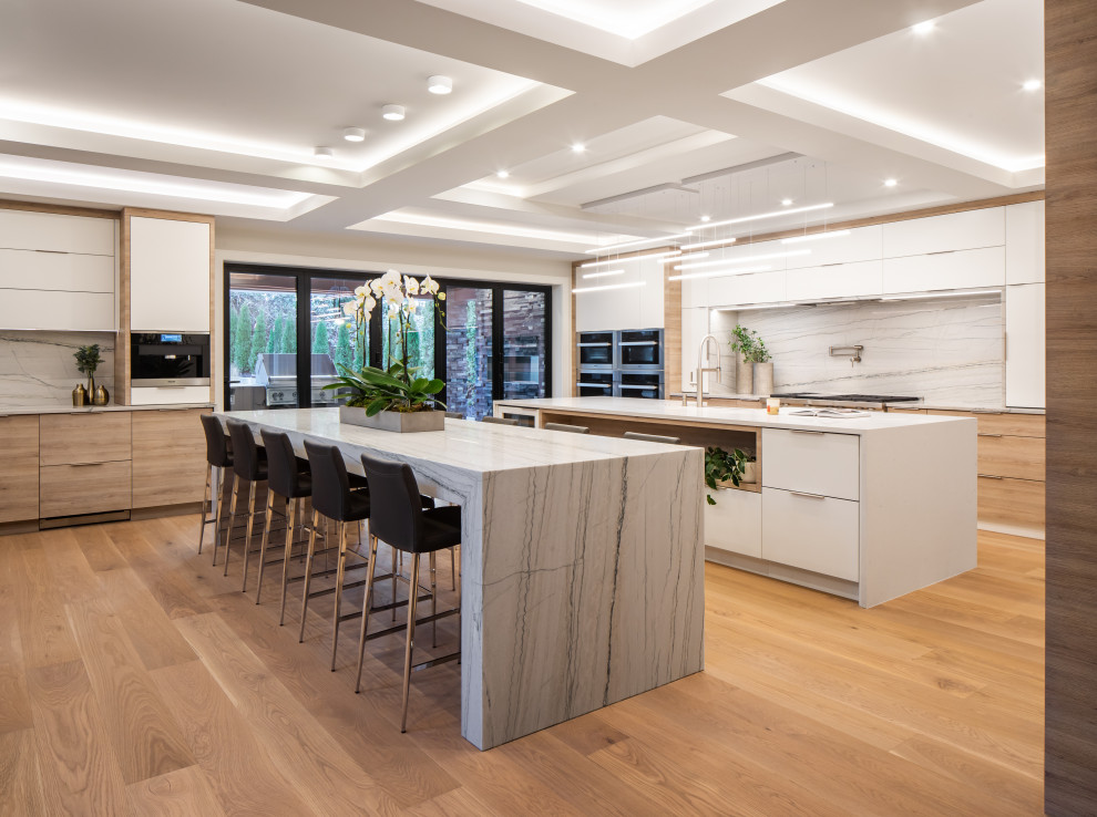 Example of a large trendy single-wall medium tone wood floor, brown floor and coffered ceiling open concept kitchen design in Vancouver with a farmhouse sink, flat-panel cabinets, light wood cabinets, quartzite countertops, white backsplash, marble backsplash, stainless steel appliances, two islands and white countertops