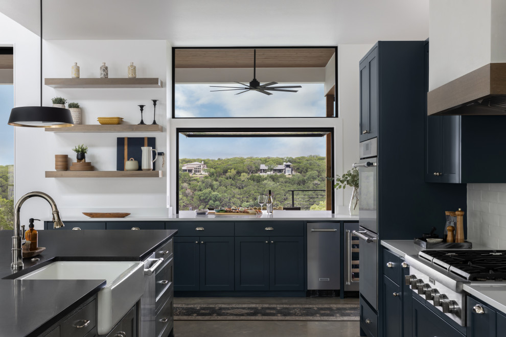 Photo of a large scandinavian l-shaped open plan kitchen in Austin with a farmhouse sink, shaker cabinets, blue cabinets, quartz benchtops, white splashback, subway tile splashback, stainless steel appliances, concrete floors, with island, beige floor, black benchtop and vaulted.