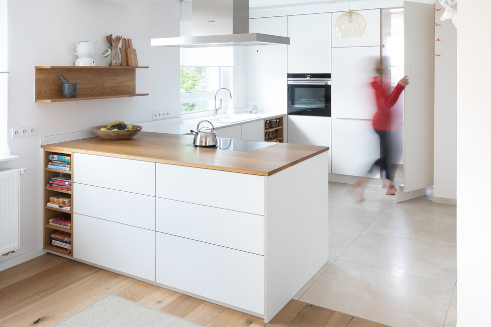 Design ideas for a mid-sized contemporary u-shaped open plan kitchen in Stuttgart with a drop-in sink, flat-panel cabinets, white cabinets, wood benchtops, white splashback, black appliances, limestone floors, beige floor, brown benchtop and a peninsula.