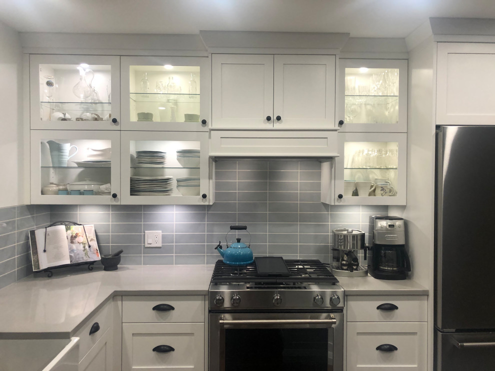 Mid-sized l-shaped eat-in kitchen photo with glass-front cabinets, white cabinets, ceramic backsplash, stainless steel appliances, an island and gray countertops
