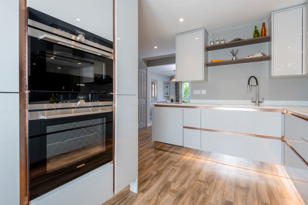 Photo of a mid-sized contemporary l-shaped open plan kitchen in Buckinghamshire with a single-bowl sink, flat-panel cabinets, grey cabinets, solid surface benchtops, metallic splashback, glass sheet splashback, stainless steel appliances, vinyl floors, no island, brown floor and white benchtop.