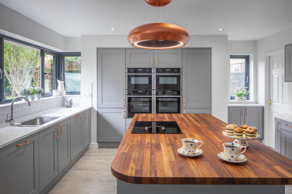 This is an example of a large transitional u-shaped eat-in kitchen in Other with an undermount sink, shaker cabinets, grey cabinets, wood benchtops, white splashback, ceramic splashback, black appliances, vinyl floors, with island, grey floor and white benchtop.