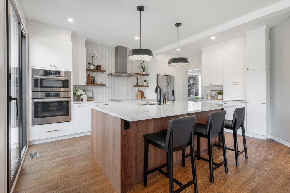 This is an example of a mid-sized midcentury l-shaped eat-in kitchen in Ottawa with an undermount sink, flat-panel cabinets, medium wood cabinets, quartz benchtops, grey splashback, ceramic splashback, stainless steel appliances, medium hardwood floors, with island and white benchtop.