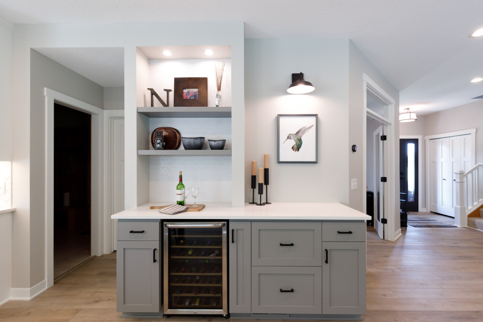 This is an example of a small transitional single-wall home bar in Minneapolis with recessed-panel cabinets, grey cabinets, quartz benchtops, white splashback, porcelain splashback, vinyl floors and white benchtop.