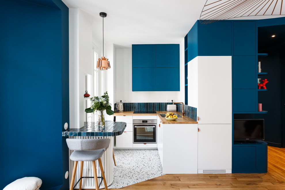 Small contemporary l-shaped open plan kitchen in Paris with a single-bowl sink, flat-panel cabinets, blue cabinets, wood benchtops, blue splashback, ceramic splashback, panelled appliances, terrazzo floors, multi-coloured floor and brown benchtop.