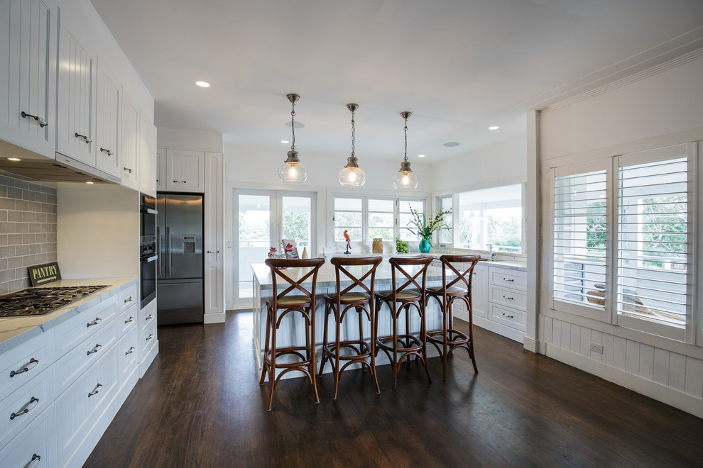 Inspiration for an expansive beach style u-shaped open plan kitchen in Sydney with a double-bowl sink, shaker cabinets, white cabinets, marble benchtops, white splashback, marble splashback, stainless steel appliances, dark hardwood floors, with island and brown floor.