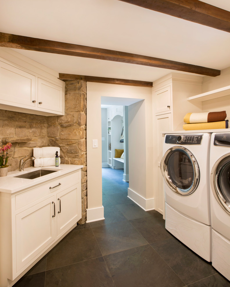 This is an example of a small country u-shaped dedicated laundry room in New York with an undermount sink, recessed-panel cabinets, white cabinets, quartz benchtops, porcelain floors, a side-by-side washer and dryer, grey floor, white benchtop, exposed beam and brick walls.