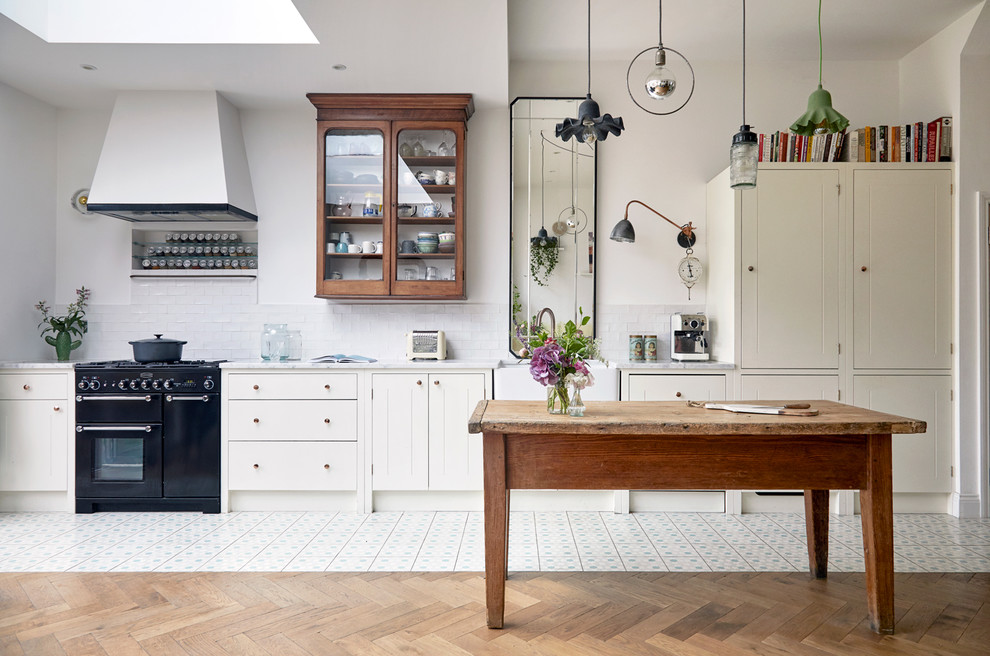 Photo of an eclectic galley kitchen in London with a farmhouse sink, flat-panel cabinets, white cabinets, white splashback, subway tile splashback, black appliances, with island, multi-coloured floor and white benchtop.
