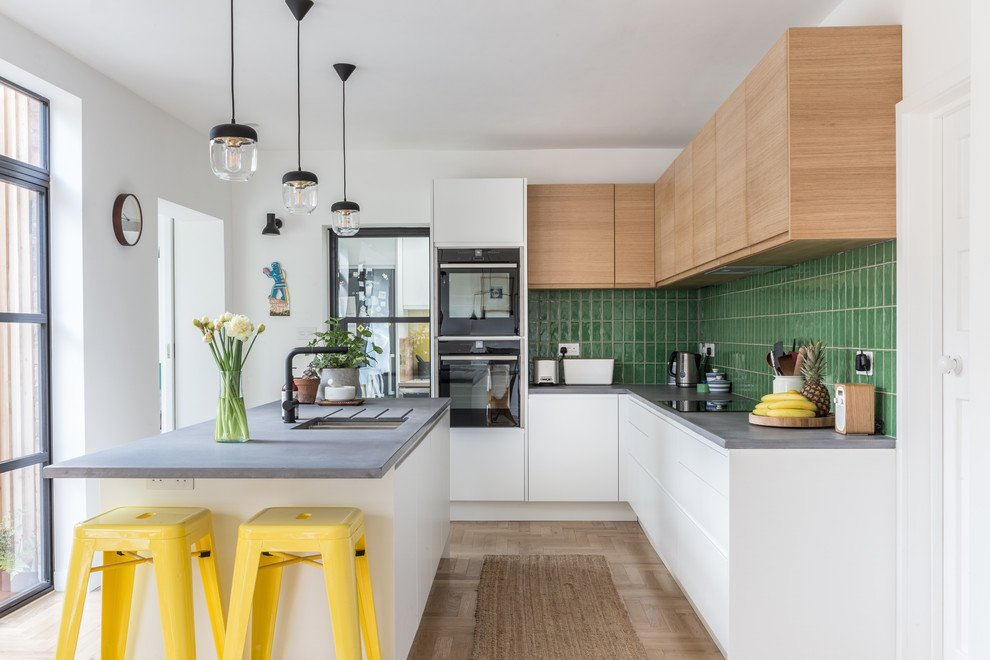 Scandinavian l-shaped kitchen in West Midlands with a double-bowl sink, flat-panel cabinets, white cabinets, green splashback, black appliances, with island, beige floor, grey benchtop and light hardwood floors.