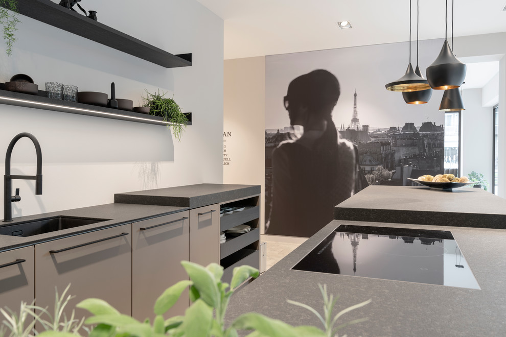 Photo of a large contemporary single-wall open plan kitchen in Cologne with a drop-in sink, flat-panel cabinets, grey cabinets, solid surface benchtops, grey splashback, black appliances, marble floors, with island, beige floor and black benchtop.