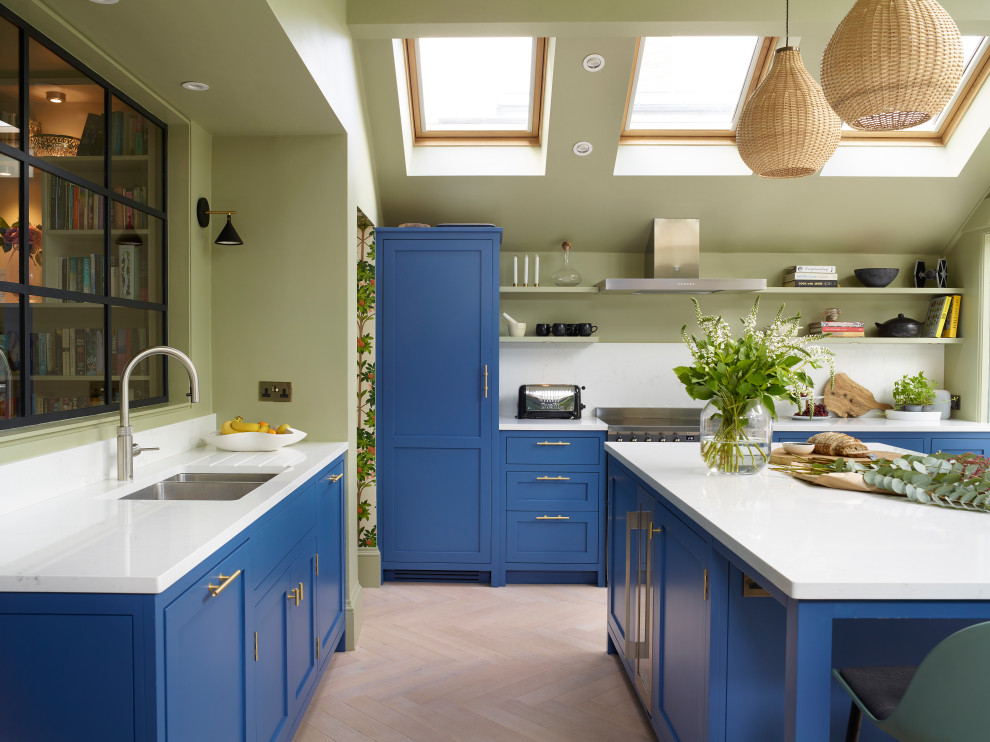 Photo of an eclectic open plan kitchen in London with a double-bowl sink, shaker cabinets, blue cabinets, composite countertops, light hardwood flooring, an island, beige floors and white worktops.