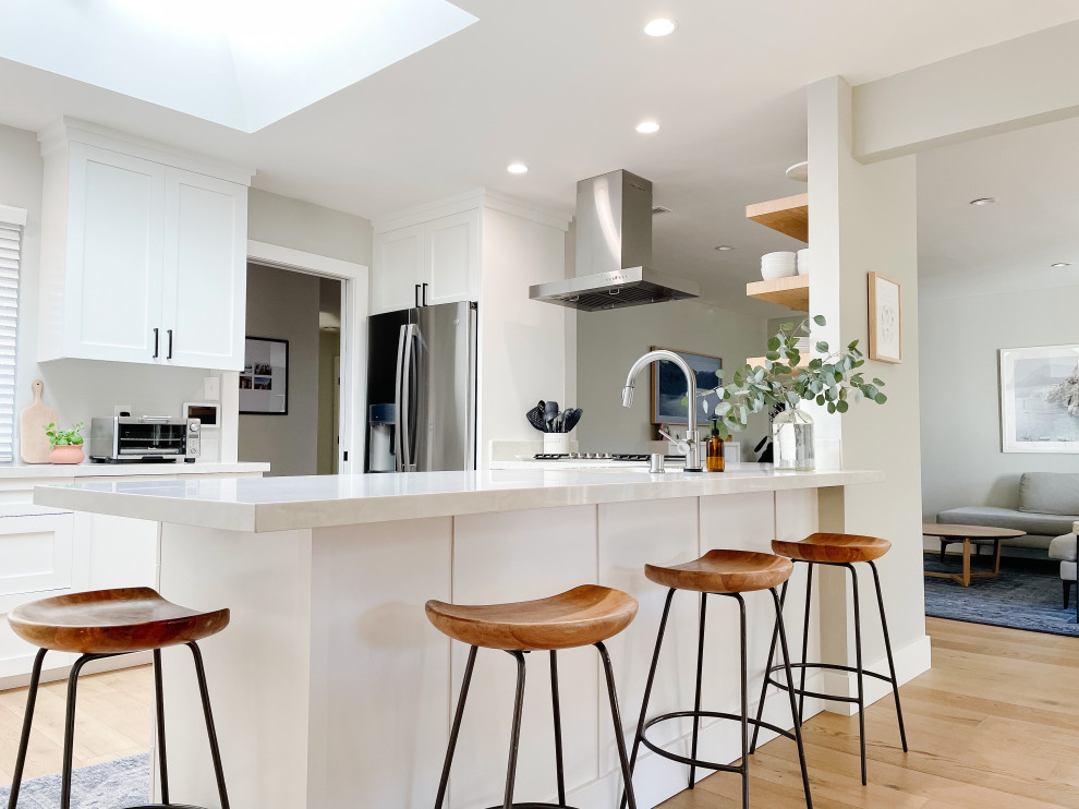 Photo of a beach style u-shaped open plan kitchen in San Francisco with an undermount sink, shaker cabinets, white cabinets, quartz benchtops, stainless steel appliances, light hardwood floors, a peninsula and grey benchtop.