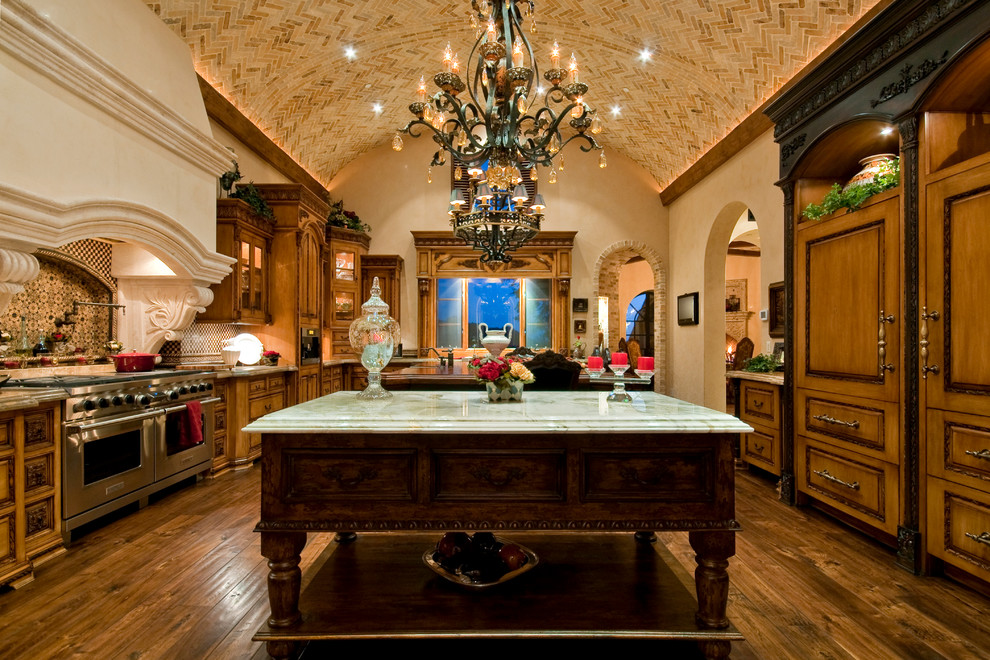 This is an example of an expansive mediterranean u-shaped open plan kitchen in Phoenix with a farmhouse sink, glass-front cabinets, distressed cabinets, glass benchtops, black splashback, marble splashback, stainless steel appliances, dark hardwood floors, multiple islands, brown floor and multi-coloured benchtop.